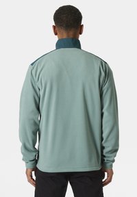 Chaqueta de forro polar en verde claro con cuello alto y acentos azules, que presenta mangas largas y una tela suave y texturada.
