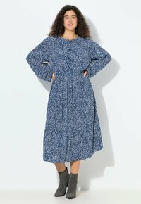 Kobieta o kręconych włosach ubrana w niebiesko-białą midi sukienkę z długim rękawem w wzory oraz szare botki, stojąca na tle białej ściany.