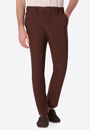 Uomo che indossa pantaloni marroni slim fit con tasche laterali e scarpe slip-on marrone scuro, in piedi con una mano in tasca.