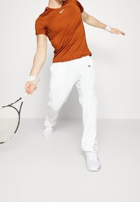 Orange sport t-shirt med en figursydd design, kombinerad med vita lösa byxor. Accessoarer inkluderar ett handledsstöd och ett tennisracket.