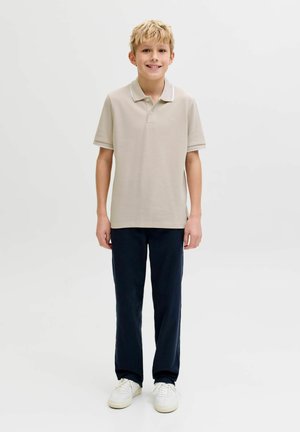 Vaaleahiuksinen poika hymyilee, seisoo rentoina käsin, pukeutuneena beigeen lyhythihaiseen poolopaitaan, tummiin housuihin ja valkoisiin tennareihin yksinkertaisella valkoisella taustalla.