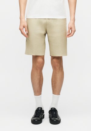 Férfi, aki bézs térdig érő rövidnadrágot, fehér zoknit, fekete tornacipőt és fehér pólót visel, egyszerű háttér előtt áll.