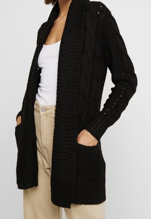 Femme portant un cardigan noir en maille torsadée ouvert avec des poches, un débardeur blanc et un pantalon beige, debout les mains dans les poches.