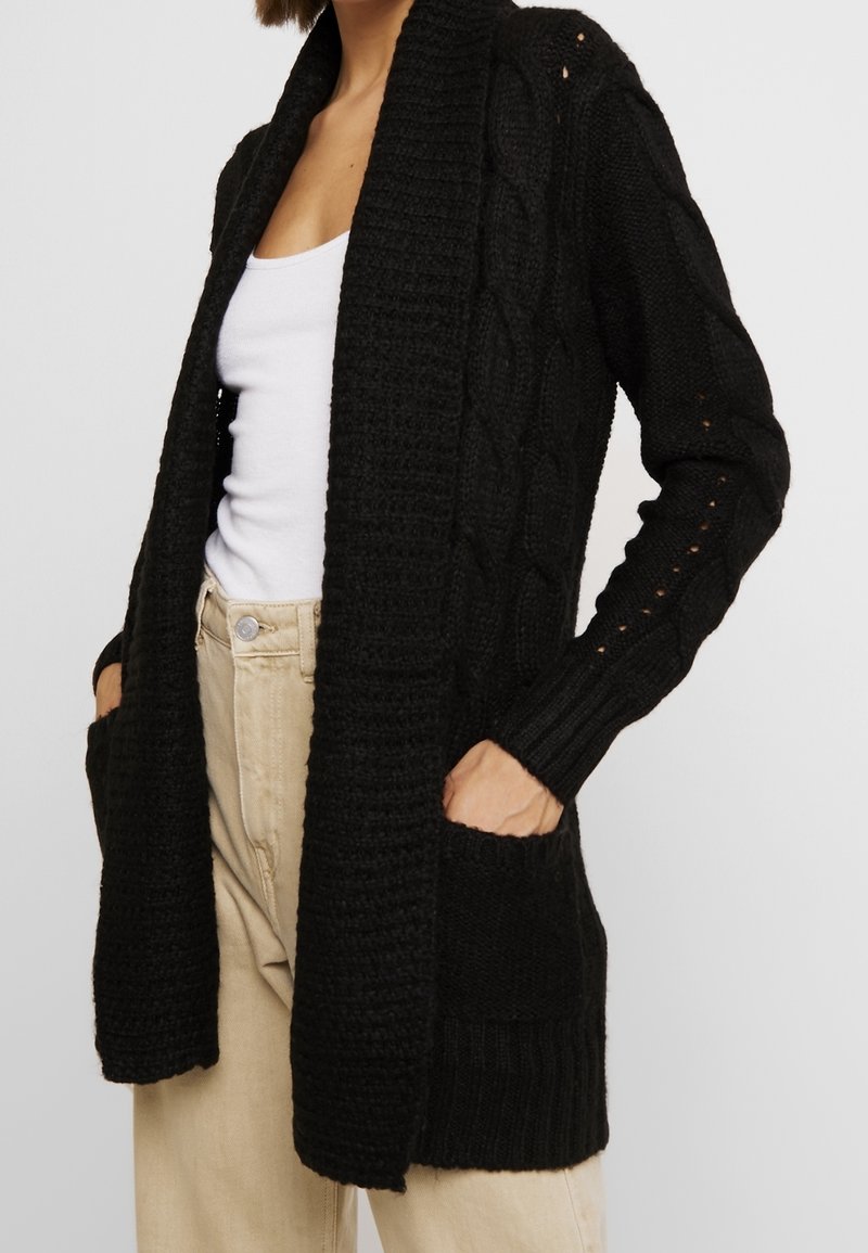 Femme portant un cardigan noir en maille torsadée ouvert avec des poches, un débardeur blanc et un pantalon beige, debout les mains dans les poches.