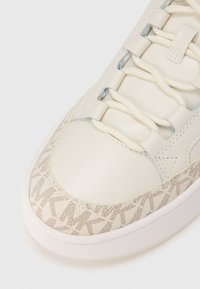 Hvid lædersneaker med et mønstret beige tekstiloverlay, rund tå og flade snørebånd. Har en glat, poleret finish og diskrete syninger.