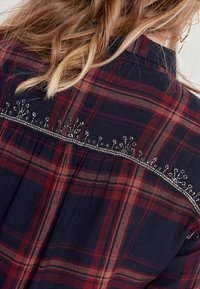 Plaid shirt with navy, red, and beige hues. Features embroidered accents along the back yoke and a soft, textured fabric.