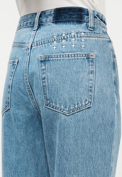 Ανοιχτό μπλε τζιν παντελόνι με ψηλή μέση, διαθέτει δύο πίσω τσέπες και λευκές ραφές σε σχήμα σταυρού στη δεξιά πλευρά.