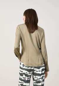 Olivenfarbene Seidenbluse mit langen Ärmeln, Rückennaht und geknöpften Manschetten, kombiniert mit schwarz-weißen gemusterten Hosen mit einem wellenförmigen Design.