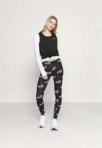 Žena stojící před jednobarevnou zdí, oblečená v černých legínách Puma s bílými logy, bílém dlouhém tričku, černém tílku a bílých teniskách.