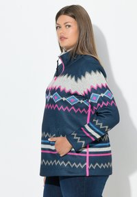 Eine Frau trägt einen marineblauen Reißverschluss-Pullover mit geometrischen Mustern in Pink, Blau, Weiß und Beige und steht vor einem weißen Hintergrund, die Hand in der Tasche.