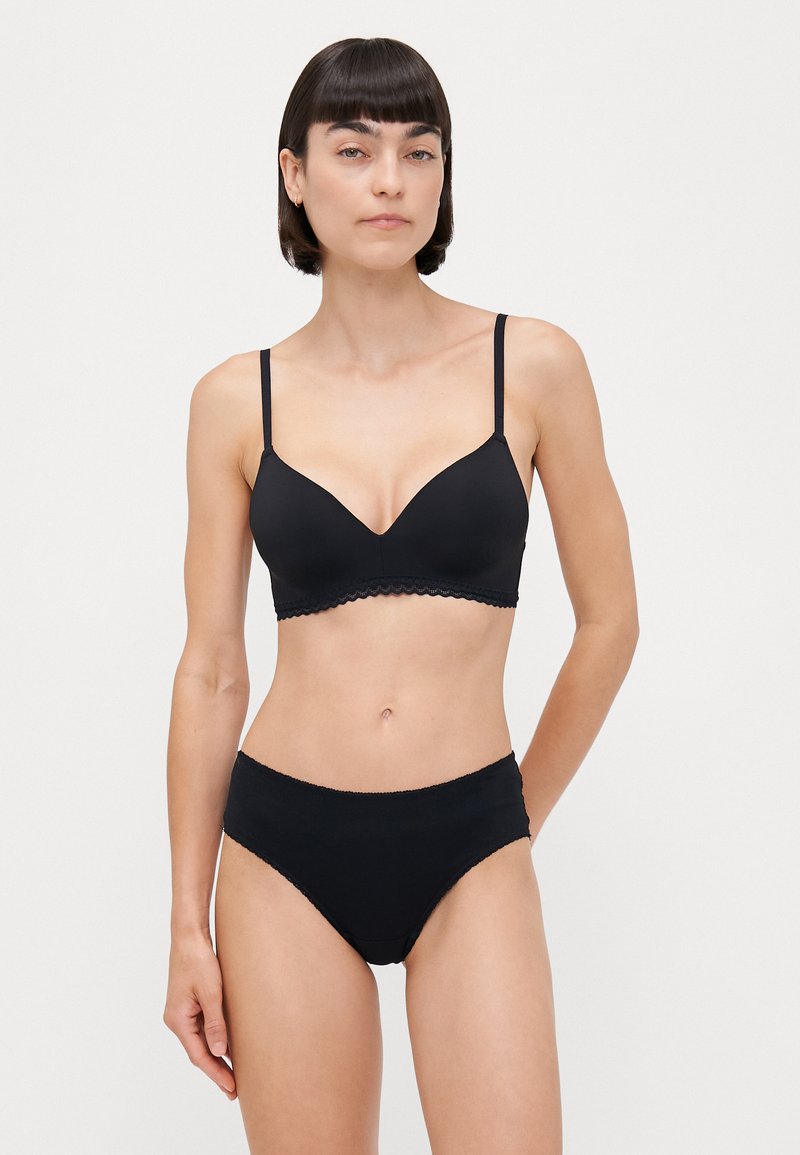 Schwarzes BH- und hochgeschnittenes Slips-Set. Der BH hat ein glattes, dreieckiges Design mit verstellbaren Trägern und gewellten Kanten. Der Slip hat eine einfarbige Optik.