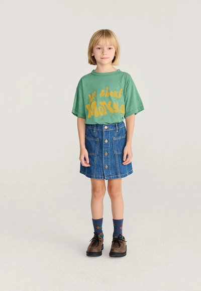 Enfant aux cheveux blonds portant un t-shirt vert avec un texte jaune, une jupe en jean boutonnée devant, des chaussettes à motifs et des chaussures marron, debout sur un fond uni.