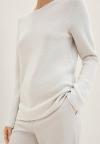 Haut gris clair à manches longues avec un col arrondi, présentant une texture côtelée subtile. Associé à un pantalon beige clair, les mains dans les poches.