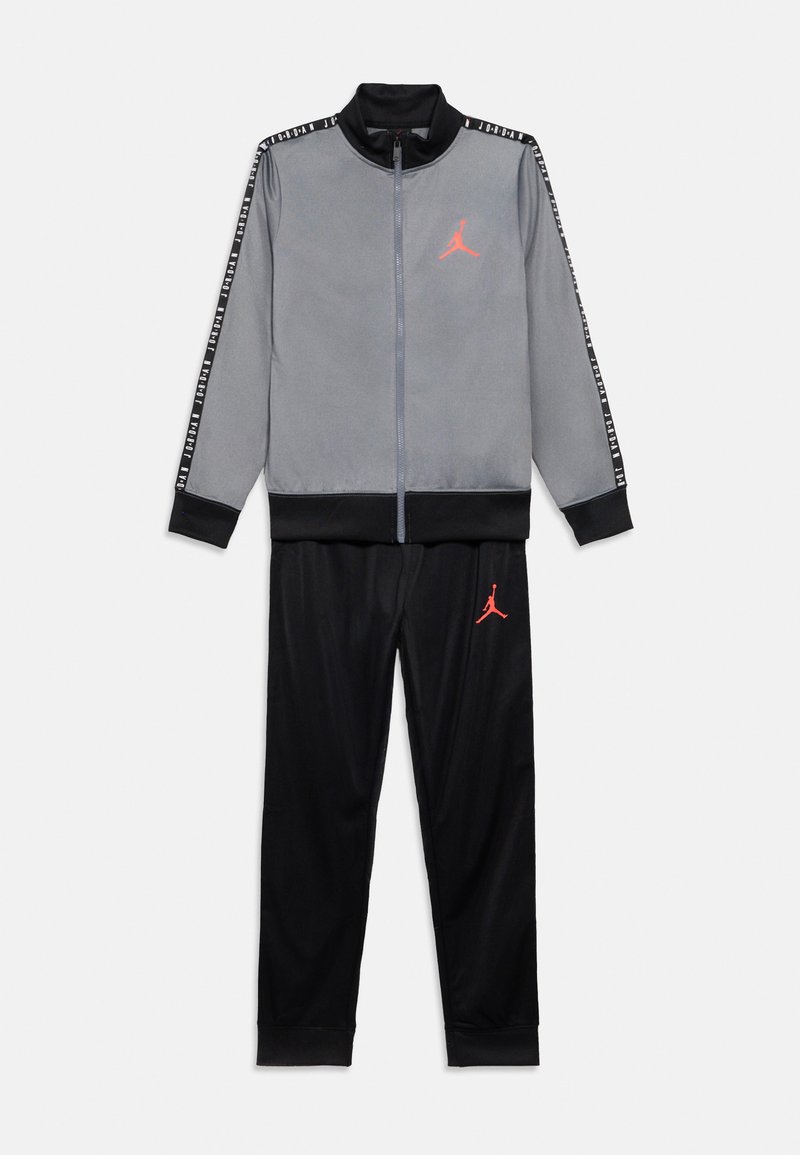 Γκρι αθλητική φόρμα που αποτελείται από ζακέτα με φερμουάρ και μαύρο γιακά, καθώς και μαύρο παντελόνι που ταιριάζει, με κόκκινα λογότυπα ως διακοσμητικά στοιχεία.