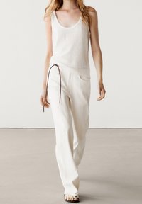 Top a fascia bianco a righe abbinato a pantaloni larghi e testurizzati con vita regolabile. Il modello indossa sandali piatti. Toni semplici e neutri.
