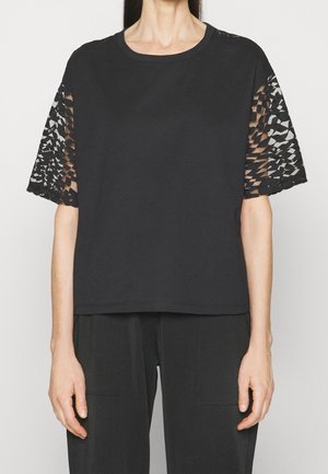 Person wearing a black short-sleeve top with geometric cutout lace sleeves and black pants against a light background.