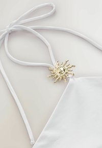 Haut dos-nu blanc avec un accent métallique en forme de soleil doré attaché à la bretelle près de l'encolure, sur un fond clair et neutre.