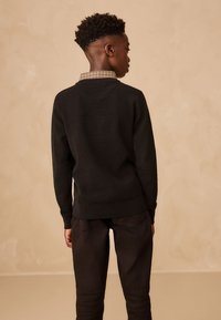 Črn pleten pulover z teksturirano površino in karo ovratnikom, kombiniran s temi jeans hlačami. Ozadje je gladke, nevtralne barve.