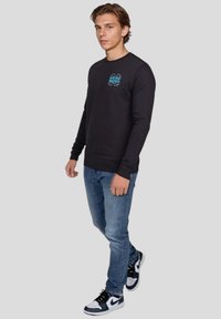 Svart långärmad tröja med rund hals, prydd med en turkos "WRONG FRIENDS" grafisk design på bröstet, i kombination med blå jeans och svarta sneakers.