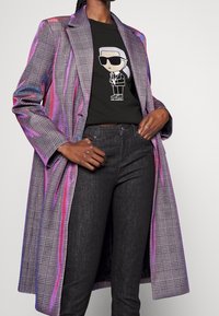 Rutigt långjacka med lila och rosa detaljer, svart grafisk T-shirt med en tecknad karaktär och mörka jeans. Tight passform, modern design.