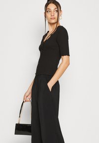 Svart ribbet cardigan med kortermet, bølger i kantene og knapper foran. Kombinert med løse svarte bukser og en liten svart håndveske.