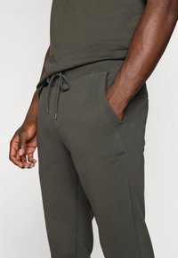 Dark green athletic pants made of textured fabric, featuring a drawstring waist and side pockets; logo printed on the leg.