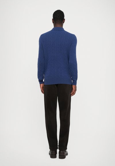 Blå cable-knit genser med krage, lange ermer, kombinert med mørke kordfløyelsbukser og mørke sko. Enkelt, teksturert design.