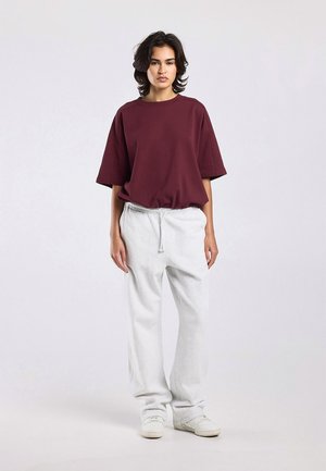 Jeune personne debout, portant un t-shirt bordeaux ample, un pantalon de survêtement blanc avec cordon et des baskets blanches sur un fond clair uni.