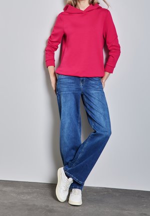Personne portant un sweat à capuche rose vif, un jean bleu à jambes larges et des baskets blanches, debout contre un mur gris les mains dans les poches.