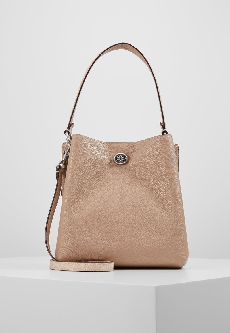 Sac à main en cuir beige texturé avec poignée supérieure, bandoulière amovible et fermeture à verrouillage tournant argenté sur une surface blanche.