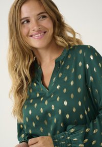 Žalia palaidinė iš lengvos medžiagos su auksinėmis taškeliais, su V formos apykakle ir raukiniais ant rankovių, suteikiančiais papildomo tekstūros.