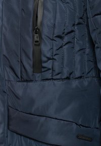 Navy blue puffer jacket fabric with quilted sections, featuring a side zipper pocket, smooth texture, and branded label.