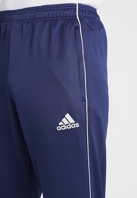 Marinblå Adidas träningsbyxor med vit piping, elastisk midja, sidoficka och Adidas-logotyp på övre lår.