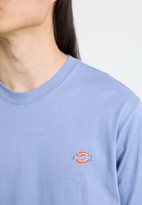 Helesinine puuvillane t-särk ümmarguse kaelusega ja väikese punase, kollase ja sinise "Dickies" logo'ga vasakul rindkere piirkonnas.