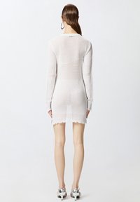 Abito bianco in maglia con maniche lunghe, con motivi testurizzati, silhouette aderente, orlo a onde e leggera trasparenza. Marchio evidente al collo.
