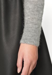 Grå stickad tröjärm med ribbade manschetter, i kombination med en slät svart lädertextur. Närbild av en hand som vilar på en mörk yta.