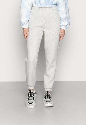 Kobieta ubrana w jasnoszare spodnie dresowe z elastycznymi mankietami i białe masywne trampki z czarnymi sznurowadłami, stojąca na tle białej, gładkiej ściany.