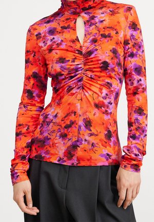 Orangenes Langarmoberteil mit lilafarbenem und schwarzem Blumenmuster, gerafftem Vorderteil, Schlüsselloch-Ausschnitt, kombiniert mit schwarzen Hosen.