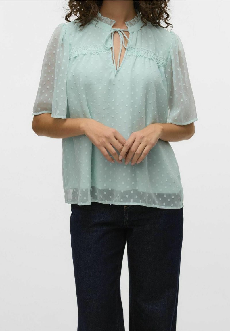 Ljus mintgrön blus med prickigt mönster, volangkant vid halsringningen, knytbar detalj och transparenta korta ärmar. Kombination med mörka jeans.