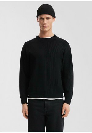 Młody mężczyzna ubrany w czarny sweter z dzianiny, czarną czapkę typu beanie, czarne spodnie oraz białą podkoszulkę, na jednolitym jasnoszarym tle.