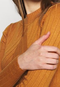 Langärmliger, gerippter Pullover in Senfgelb mit hohem Kragen und strukturiertem Muster. Nahaufnahme eines Arms mit einer ruhenden Hand.