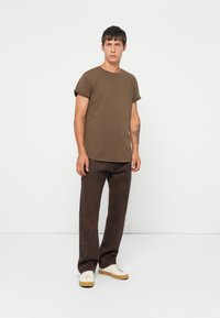 Brun kortermet t-skjorte med rund hals, kombinert med mørkebrune bukser og hvite sneakers med beige såler, stående mot en hvit bakgrunn.