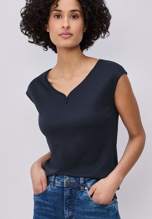 Frau mit lockigem Haar, die ein dunkles, ärmelloses Oberteil mit zwei Knöpfen trägt, die Hände in den Taschen einer blauen Jeans, steht vor einem schlichten Hintergrund.