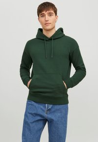 Felpa con cappuccio verde scuro, dotata di tasca frontale, dettagli con cordino e polsini a costine, abbinata a jeans azzurri chiari.