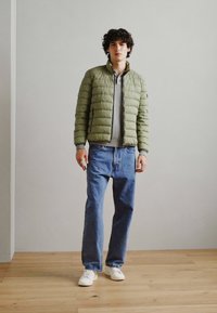 Giubbotto a vento di colore verde chiaro, in tessuto testurizzato, abbinato a una felpa grigia e jeans blu. Il modello indossa sneakers bianche e si trova su un pavimento in legno.