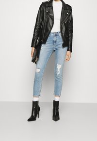 Chaqueta de moto de cuero negra, top blanco de canalé, vaqueros cortos de mezclilla azul claro desgastados y botines de tacón negros. Fondo simple.