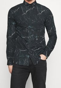 Man in een zwart slim-fit shirt met een wit en blauw marmerachtig patroon, gecombineerd met zwarte broek, met één hand in de zak.