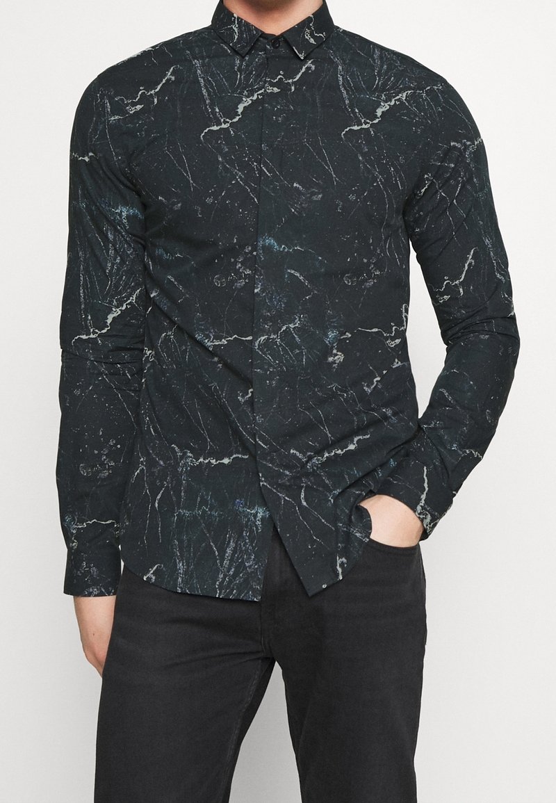 Man in een zwart slim-fit shirt met een wit en blauw marmerachtig patroon, gecombineerd met zwarte broek, met één hand in de zak.