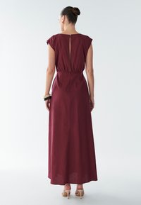 Maxi šaty vínovej farby s krátkymi rukávmi, zošitý pás a detail otvorených chrbta. Hladký materiál s jemným leskom. Oblečené s béžovými sandálmi na remienky.