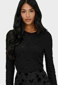 Donna con lunghi capelli neri indossa un top a maniche lunghe di tessuto nero e una gonna nera con motivo floreale, rivolta in avanti su uno sfondo neutro.